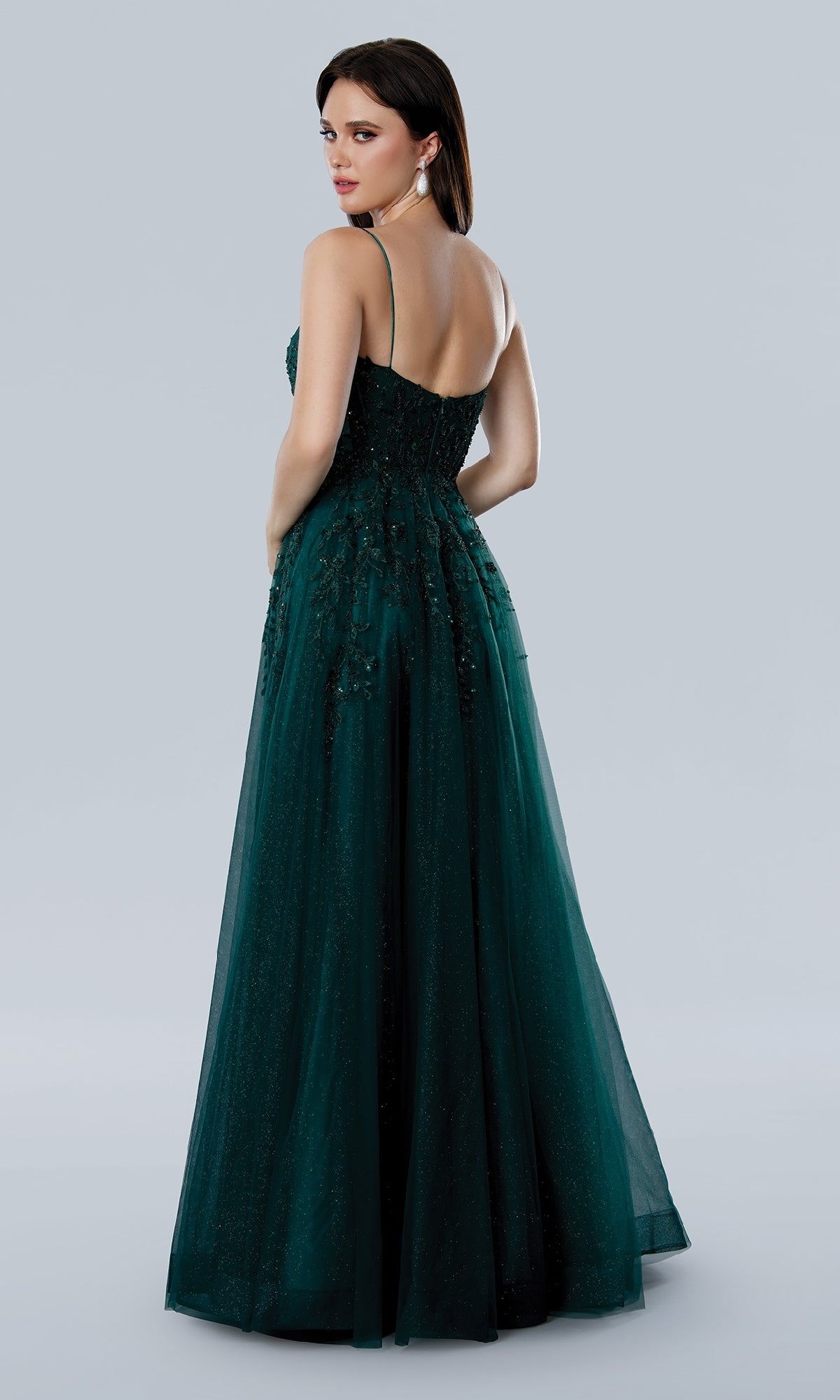 Woman wearing the Stella Couture 25109 long dark green prom dress with spaghetti straps, low back, and floral embellishments, standing against a plain light gray background.